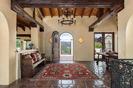 Custom wrought iron chandelier and sconces illuminating a warm, rustic entryway with wooden beams and a patterned rug.