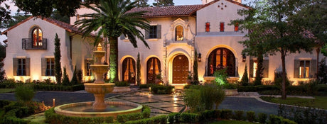 Custom wrought iron outdoor lighting fixtures illuminating the exterior of a luxurious home with a fountain and lush landscaping.