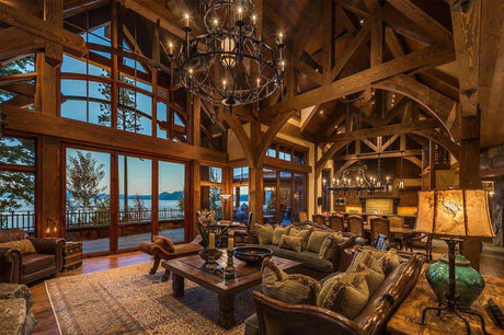 Luxurious living room featuring a custom wrought iron chandelier, exposed wooden beams, and large windows framing a tranquil lakeside view.