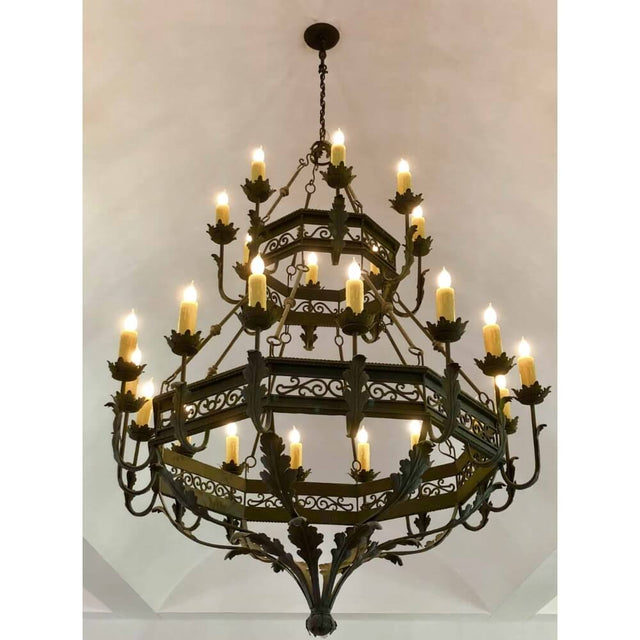 An ornate wrought iron chandelier featuring multiple tiers and elegantly designed candle holders. The fixture is adorned with intricate leaf motifs and emits a warm glow from its lit candles, creating a welcoming ambiance.