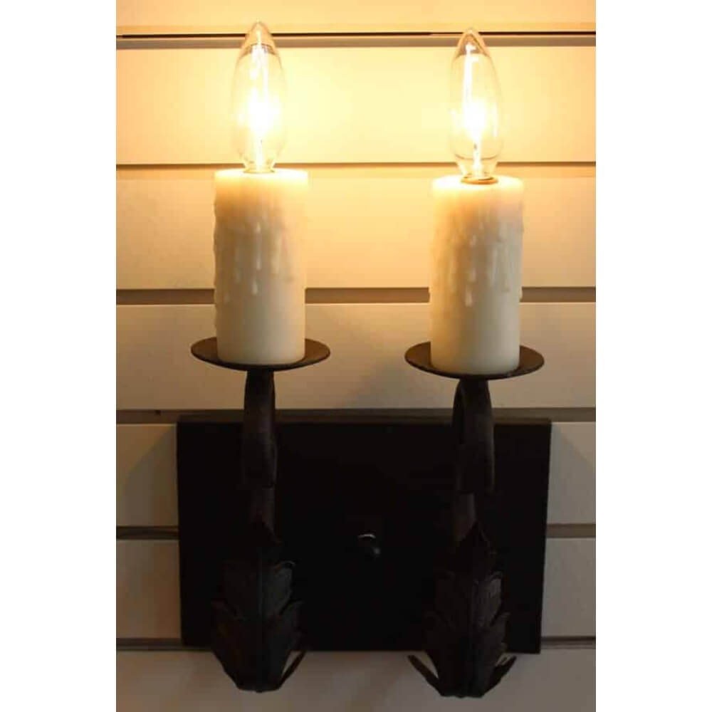 A pair of elegantly designed wrought iron sconces featuring two candle-like lights atop decorative holders. The candles, resembling wax with dripped edges, emit a warm glow, enhancing the ambiance of any space. The intricate ironwork showcases an artistic leaf motif, adding a touch of rustic charm to the fixture.