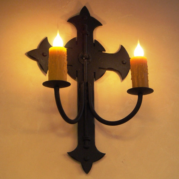 A decorative wrought iron wall sconce featuring a cross design and two candle holders. Each holder displays a rustic, textured yellow candle that emits a warm glow, enhancing a cozy atmosphere.