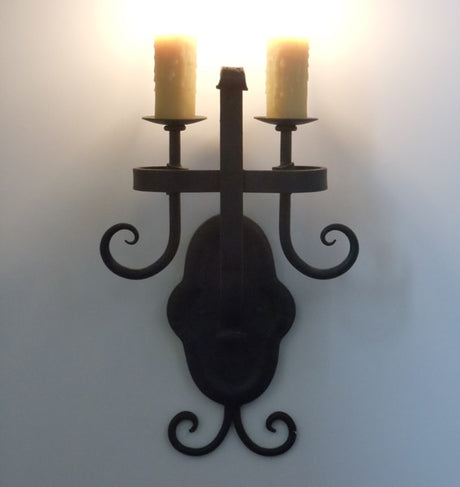 A decorative wall sconce featuring two candle holders with a refined, curved design. The fixture has an ornate backplate and is finished in a dark metal, adding elegance and warmth to any space.