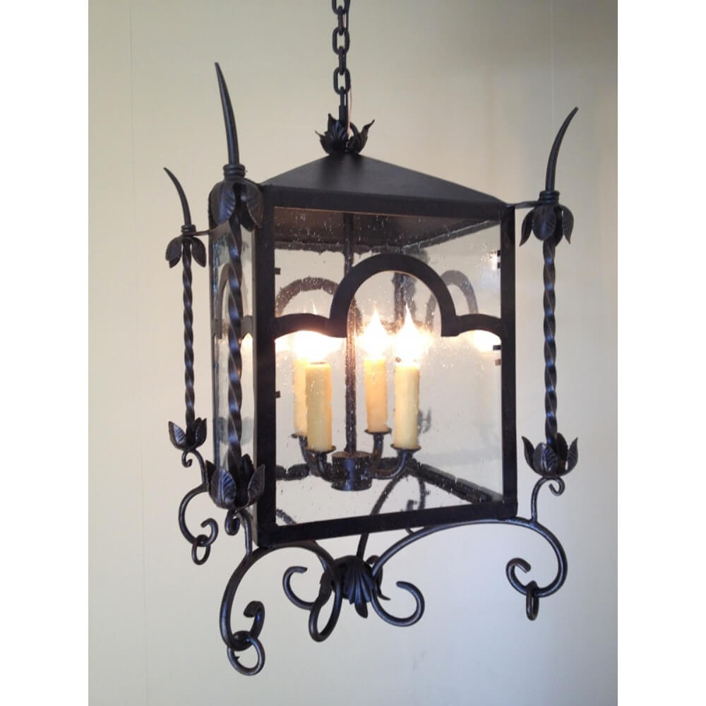 A decorative wrought iron pendant featuring a square frame with curved accents, glass panels, and ornate leaf motifs. It holds four candle-like lights, enhancing any space with a warm, inviting glow.