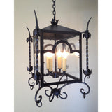 A decorative wrought iron pendant featuring a square frame with curved accents, glass panels, and ornate leaf motifs. It holds four candle-like lights, enhancing any space with a warm, inviting glow.