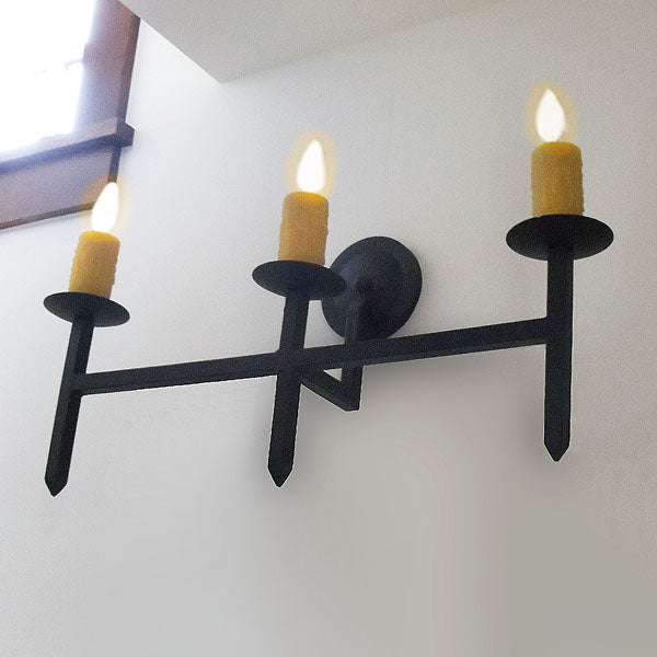 A three-armed wall sconce featuring a sleek, dark iron frame with yellow candle-like lights. The fixture rests against a light-colored wall, creating a warm, inviting atmosphere.