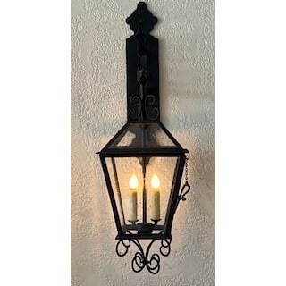 A decorative wall-mounted sconce featuring a geometric glass design with intricate scrollwork. Two candle-like bulbs illuminate the fixture, evoking a warm, inviting ambiance. The textured black finish adds a touch of elegance and charm to any space.