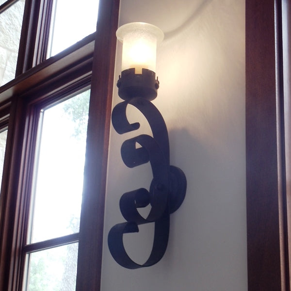 A decorative wall sconce featuring elegant twisted wrought iron accents. It supports a frosted glass shade that casts a warm glow, enhancing interior spaces with its artistic design.