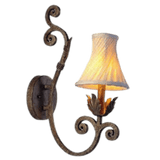 A decorative wall sconce featuring an ornate wrought iron design with elegant curves. It showcases a soft, pleated shade that emits warm light, complemented by leafy accents at the base, adding a touch of classic charm to any space.