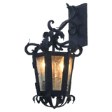 A decorative wall sconce featuring intricate wrought iron detailing, lantern-style design, and a warm glow emanating from three light bulbs.