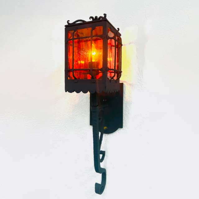 A decorative wrought iron wall sconce featuring a vibrant orange glass lantern. The intricate metalwork surrounds the vivid light source, casting warm illumination against the wall.