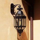 A decorative wall sconce featuring intricate wrought ironwork and a glass enclosure. Its elegant shape and detailed design make it a charming accent for outdoor or indoor spaces, enhancing the ambiance with a warm glow.