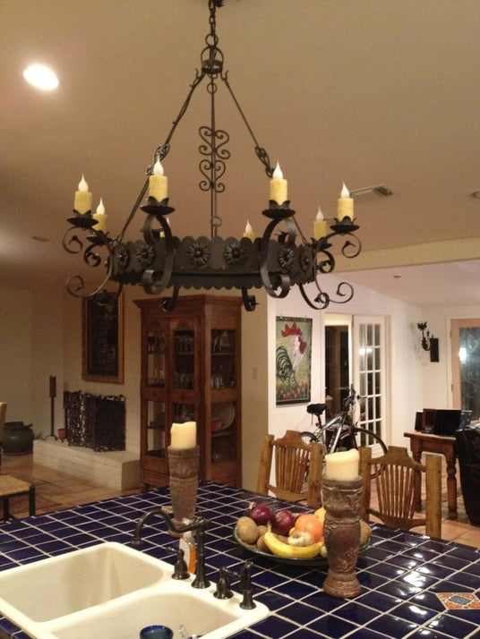 A beautifully crafted wrought iron chandelier with decorative flourishes hangs above a blue tiled kitchen island, featuring six candle-like lights. The warm ambiance is complemented by wooden furnishings and a vibrant fruit bowl, creating an inviting atmosphere.