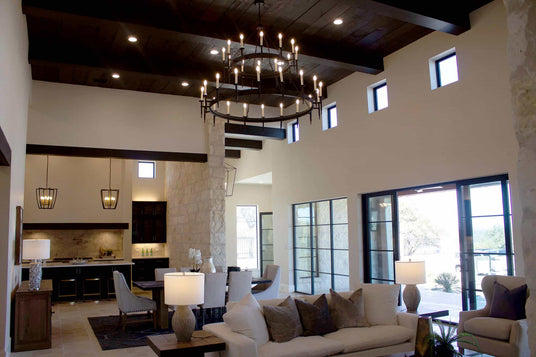 A spacious living area featuring a large wrought iron chandelier with multiple candle-like lights suspended from a dark wooden ceiling. Elegant pendant fixtures hang above a kitchen area with modern cabinetry, while natural light pours in through expansive glass doors and windows, illuminating the stylish furnishings and decor throughout the space.