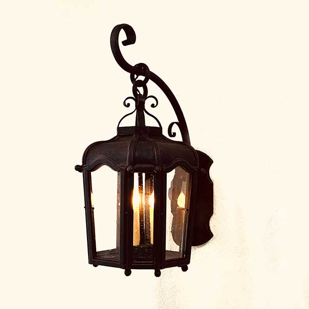 A decorative wrought iron wall sconce featuring a rounded top, curved design, and a warm glow from exposed bulbs inside. The intricate detailing and elegant hook enhance its charm.