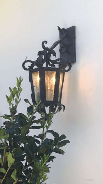 A decorative wall sconce featuring intricate wrought iron detailing, lantern-style design, and a warm glow emanating from three light bulbs. The fixture is mounted on a white wall, surrounded by lush greenery, enhancing its elegant and inviting look.