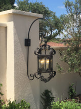 A beautifully crafted outdoor sconce featuring ornate wrought iron detailing and a lantern-style glass enclosure, illuminating a lush garden setting.