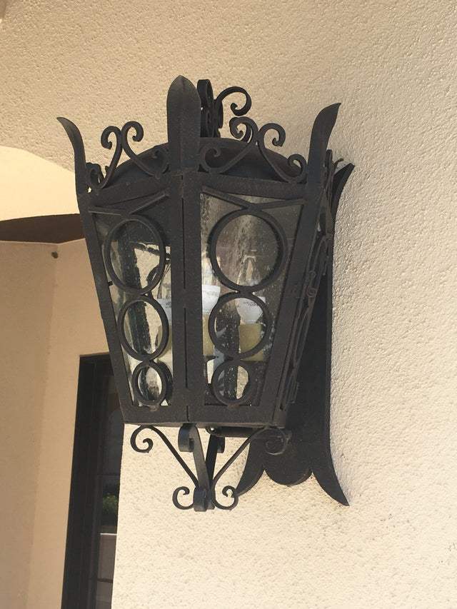 A decorative wrought iron sconce featuring intricate scrollwork and circular glass panels, mounted on a textured wall. The design combines elegance with rustic charm, perfect for illuminating outdoor spaces.