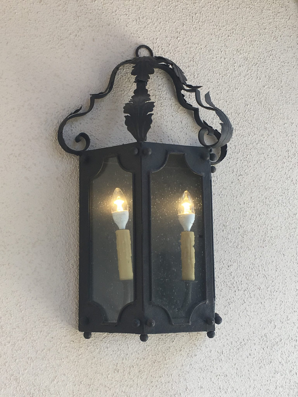 A beautifully crafted wrought iron sconce featuring intricate scrollwork and a decorative top. Dual glass panels enclose two glowing bulbs, enhancing its classic elegance.