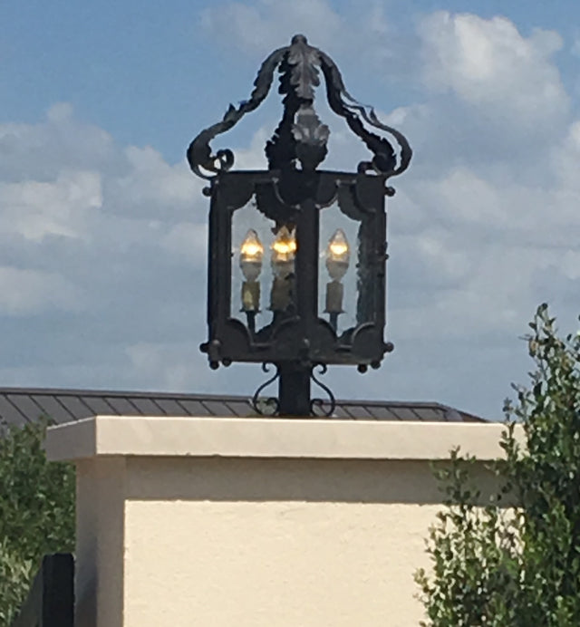 A decorative wrought iron sconce featuring intricate scrollwork and a glass enclosure, showcasing four warm light bulbs. The fixture sits atop a stucco wall with a clear blue sky in the background, enhancing its elegant design.