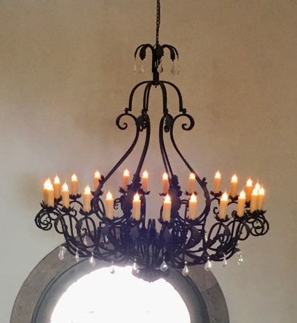 A beautifully crafted wrought iron chandelier with multiple elegant arms, adorned with warm, flickering candle-like lights and delicate crystal accents, suspended from a chain against a light, neutral background.