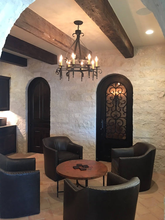 A beautifully crafted wrought iron chandelier hangs from a wooden beam ceiling in a cozy space. Its design features elegant curves and candle-like lights that enhance the warm atmosphere. The room includes dark wooden doors, an intricately designed iron door, and plush armchairs surrounding a rustic round table, creating a welcoming setting for relaxation and conversation.