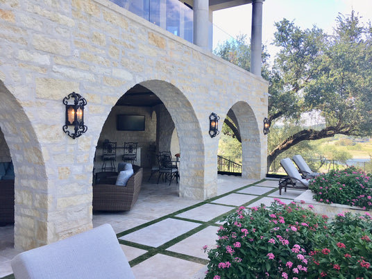 Elegant wrought iron sconces illuminate a charming outdoor space with arched stone walls. Lush greenery and pink flowers enhance the inviting atmosphere.