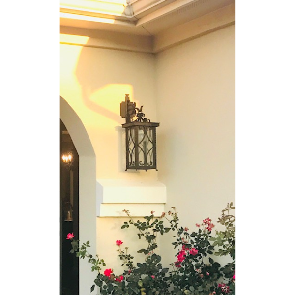 A beautifully crafted wrought iron sconce, featuring intricate scrollwork and glass panels, mounted on a light-colored wall. Vibrant flowers in the foreground enhance the aesthetic, casting soft evening shadows.