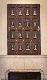 A vintage, ornately carved wooden panel featuring small, detailed figurines in recessed frames, mounted above a decorative stone fireplace.