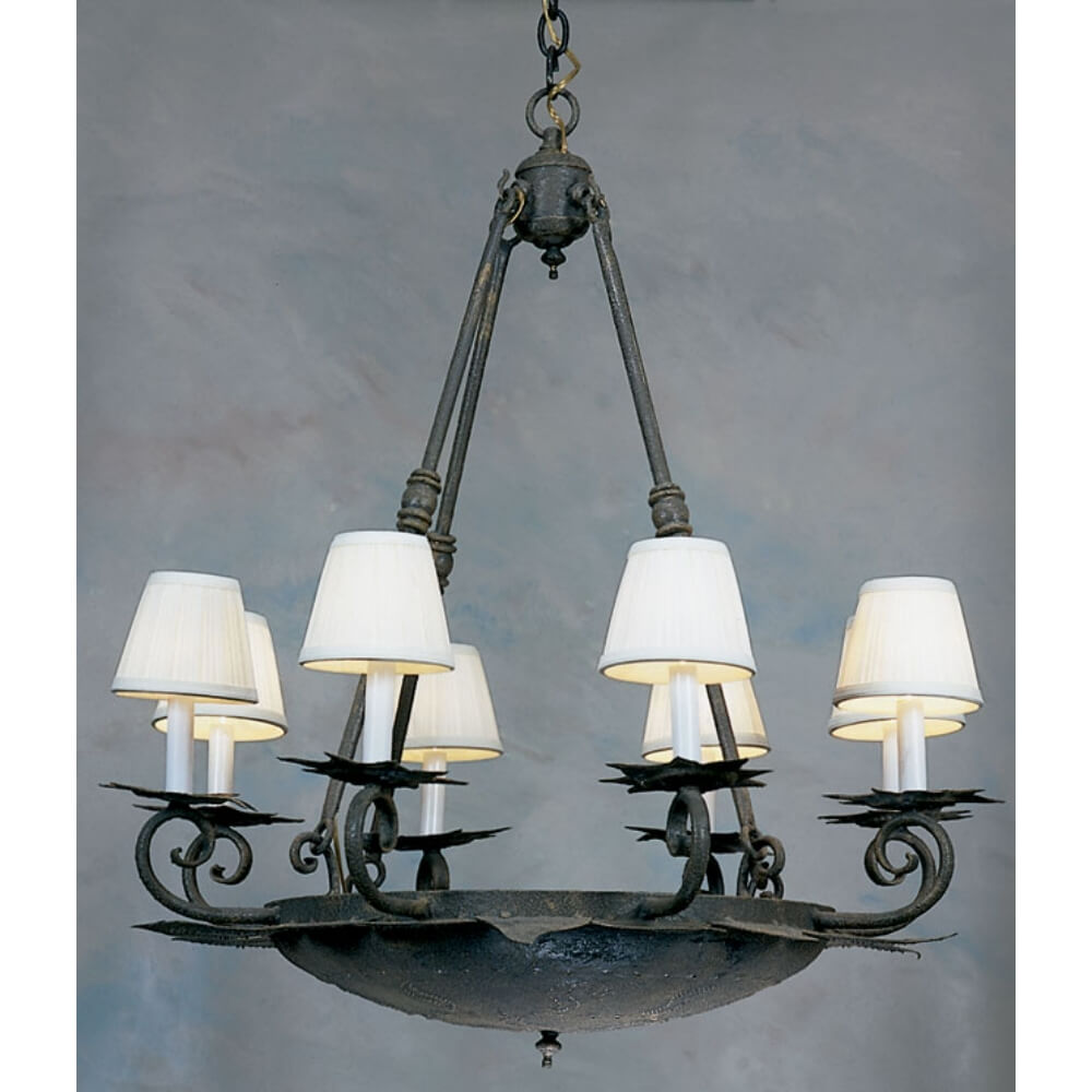 An elegant wrought iron chandelier features a round base with a textured, dark finish, adorned with six candle-style lights topped with white fabric shades. The design includes intricate scrollwork and a chain for hanging, creating a warm and inviting atmosphere.