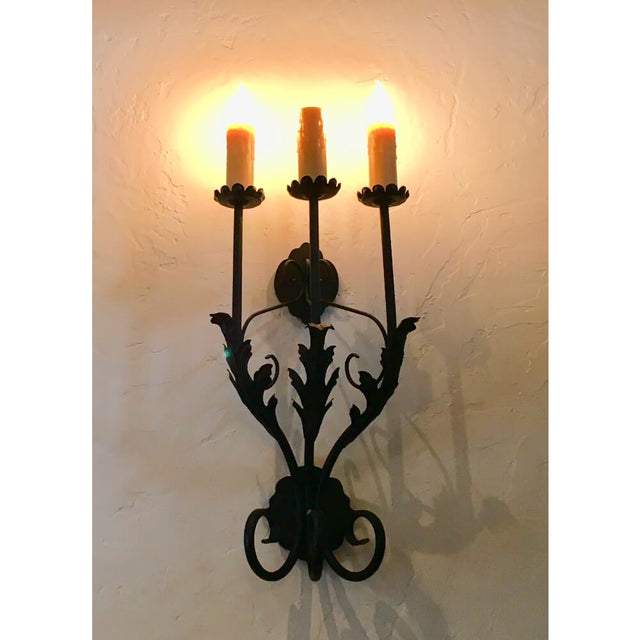 A decorative wall sconce featuring three candle-like bulbs, elegantly designed with curling leaves and intricate metalwork, casting warm light against a textured wall.