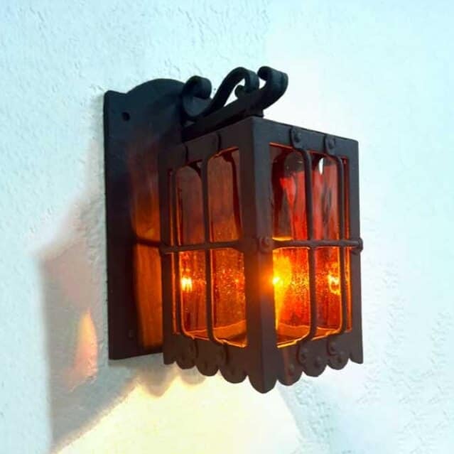 A wrought iron wall sconce featuring a square design with vibrant amber glass panels. The intricate metalwork includes decorative scrolls and a sturdy mount, casting a warm glow.