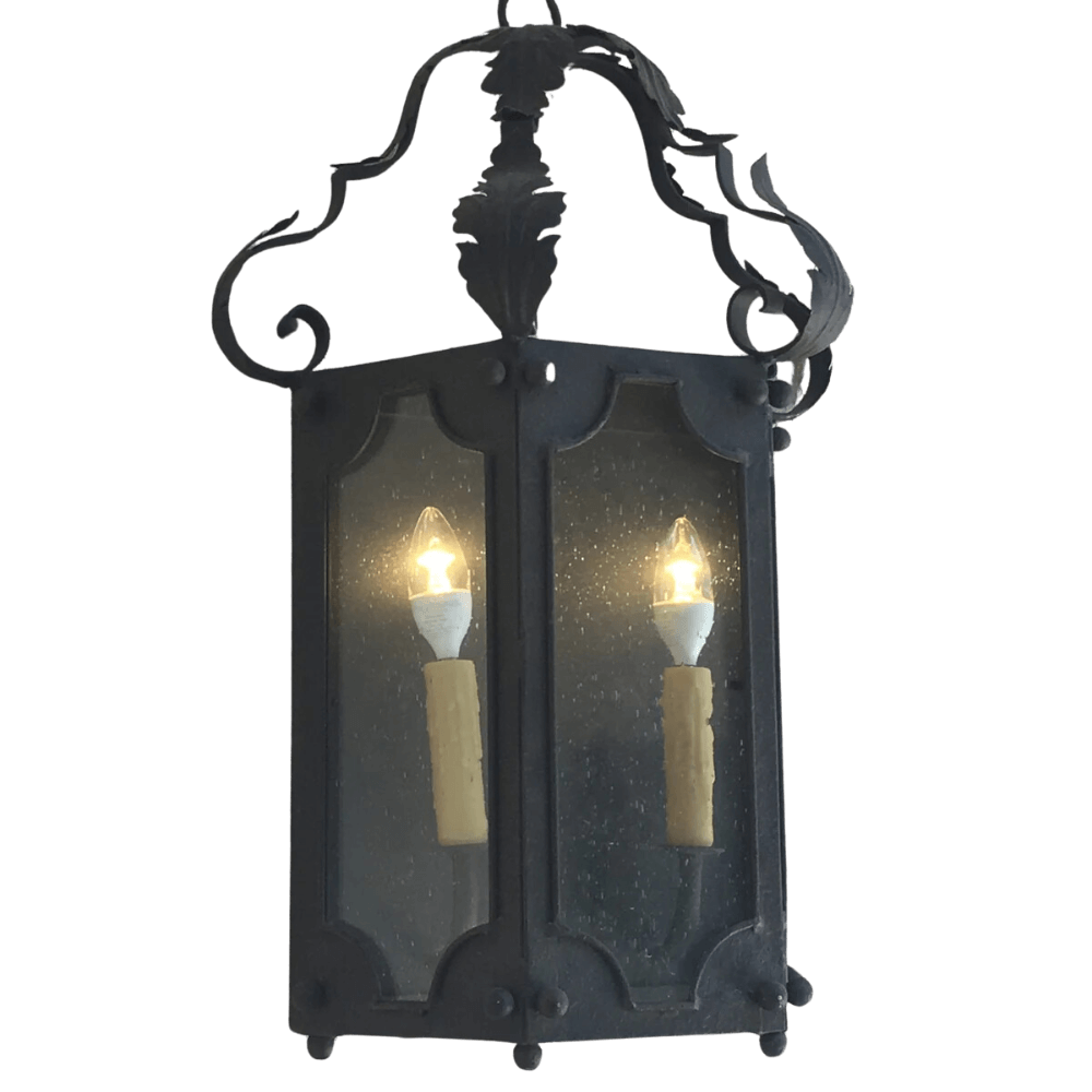 A hand-crafted wrought iron pendant featuring two illuminated candle-like bulbs, elegantly encased in glass panels with decorative curves and a classic design.