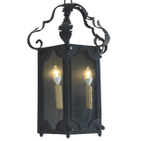 A hand-crafted wrought iron pendant featuring two illuminated candle-like bulbs, elegantly encased in glass panels with decorative curves and a classic design.