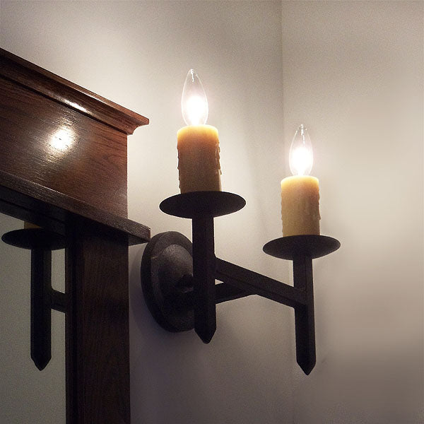 A handcrafted wrought iron wall sconce featuring two candle-like lights. The fixture showcases a rustic design with a dark finish, adding warmth and elegance to any space.