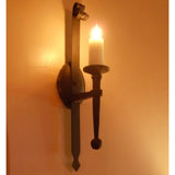 A wrought iron wall sconce featuring a slender, elongated design. It supports a white candle, casting a warm glow against the textured wall, highlighting the elegant craftsmanship and vintage appeal.