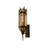 A sleek wrought iron wall sconce featuring a cylindrical glass shade with decorative ironwork. The design incorporates a pointed base and intricate detailing, creating an elegant illumination effect suitable for diverse interiors.