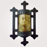 A decorative wall sconce featuring intricate wrought iron detailing and a cylindrical amber glass center. The design showcases elegant cutouts and ornate accents, creating a warm, inviting glow for any space.