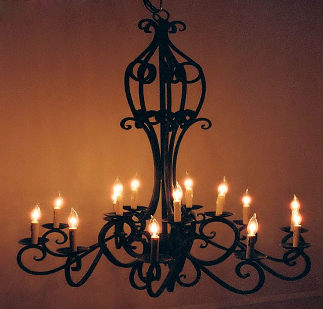 A large wrought iron chandelier featuring an intricate design with curling arms, holding multiple candle-like lights that emit a warm glow against a dark background.