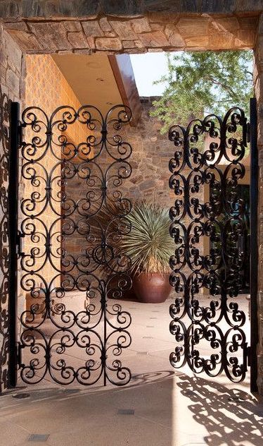 A pair of ornate wrought iron gates featuring intricate scrollwork opens into a patio area adorned with stone walls and desert plants.