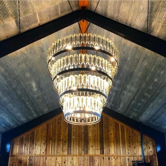 A stunning chandelier features multiple tiers of glass elements, elegantly surrounded by wrought iron rings. The fixture is suspended from a rustic ceiling, casting a warm glow in a wooden interior space.