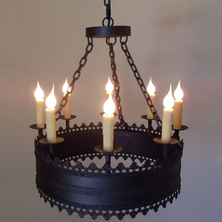 A bold wrought iron chandelier featuring elegant candle-like lights, adorned with intricate decorative detailing along the edge. The design highlights a rustic charm, perfect for enhancing warm, inviting spaces.