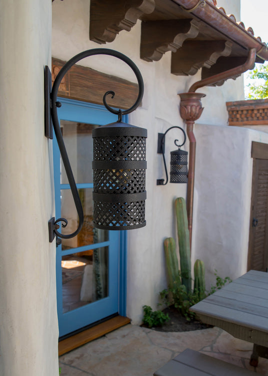 A black wrought iron sconce with intricate cutout designs hangs by a blue door, complementing the textured wall and surrounding greenery.