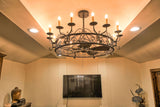 A wrought iron fandelier—a combination of a chandelier and ceiling fan—hangs from the ceiling, featuring an intricate scrollwork design. The circular frame supports multiple candle-style lights with faux wax-drip details, casting a warm glow. Beneath the ornate structure, a discreet ceiling fan with dark wooden blades blends seamlessly, providing both elegance and functionality.