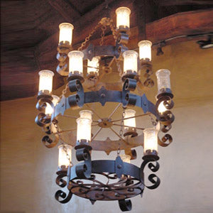 A large, ornate chandelier featuring multiple candle-like glass shades atop a circular wrought iron frame. The design showcases elegant curves and intricate scroll work, enhancing the warm ambiance in any space.