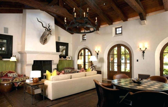 A spacious living room featuring a custom wrought iron chandelier with elegant candles, wooden beamed ceilings, arched windows, and rich textiles that enhance the warm ambiance.