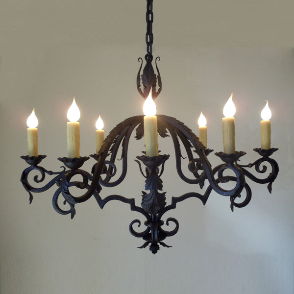 A classic wrought iron chandelier featuring intricate scrollwork and floral motifs. The design includes six candle-like lights with warm, amber bulbs, suspended from a decorative chain. Ideal for adding elegance to living spaces.