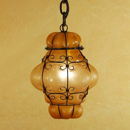 A decorative pendant light featuring a warm, amber glass shade enclosed in intricate wrought iron detailing. The design showcases elegant curves and loops, enhancing a cozy, vintage aesthetic.