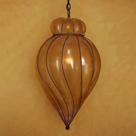 A beautifully crafted pendant light featuring a curved, elongated glass body with vertical lines, topped with a rounded finial. The warm, amber hue creates a soft, inviting glow, perfectly complemented by the intricate wrought iron detailing.