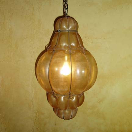 A decorative pendant light featuring a textured, amber glass shade encased in wrought iron, suspended from a chain against a warm, earthy wall.